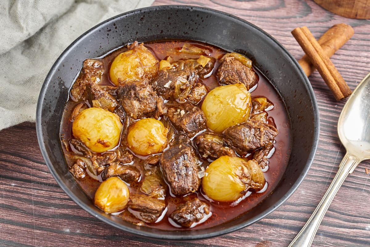 greek stifado with beef and onion in a plate with a spoon and cinnamon sticks next to it