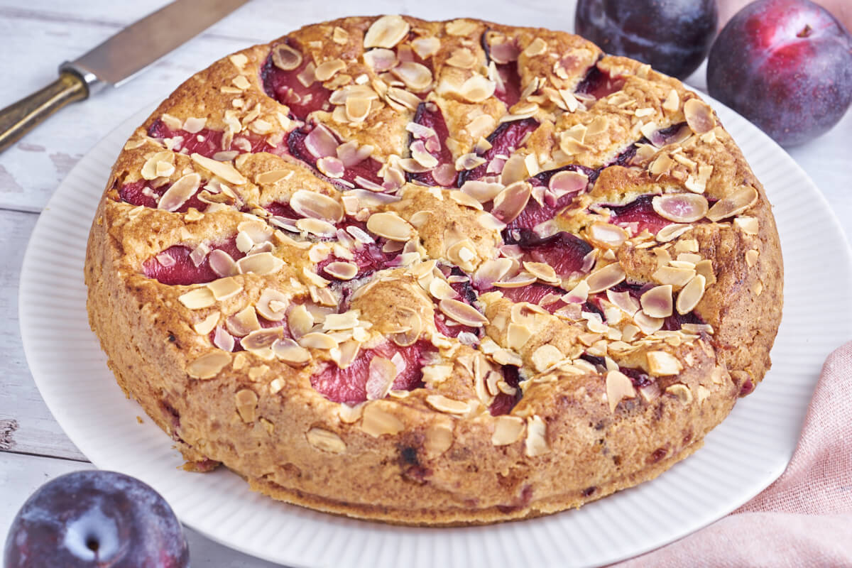 cake with plums and almond flakes on a white platter with fresh plums and a knife