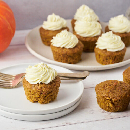 Pumpkin muffins with cream cheese frosting on a small cake plate with a platter of more cupcakes in the background