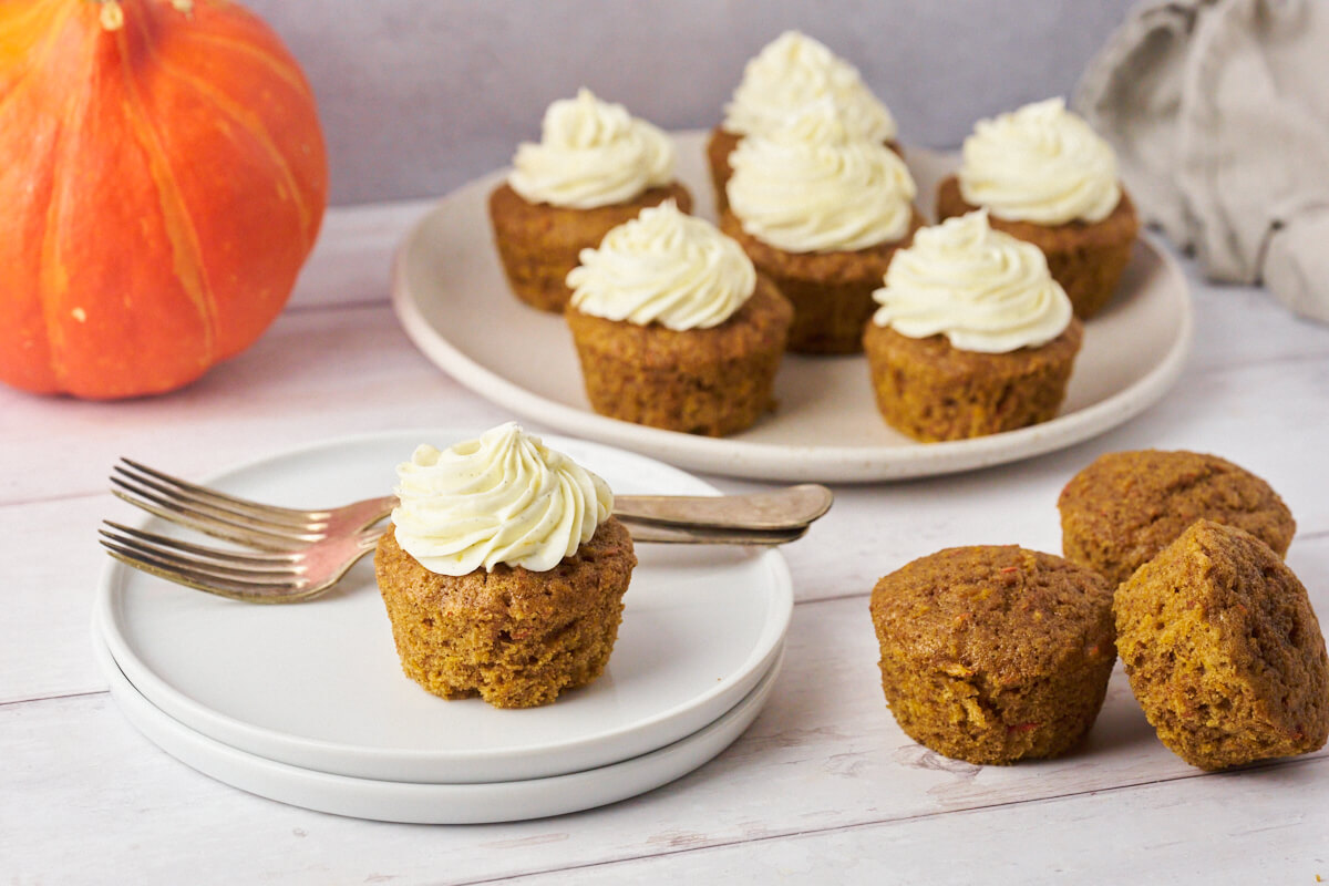 Pumpkin muffins with cream cheese frosting on a small cake plate with a platter of more cupcakes in the background