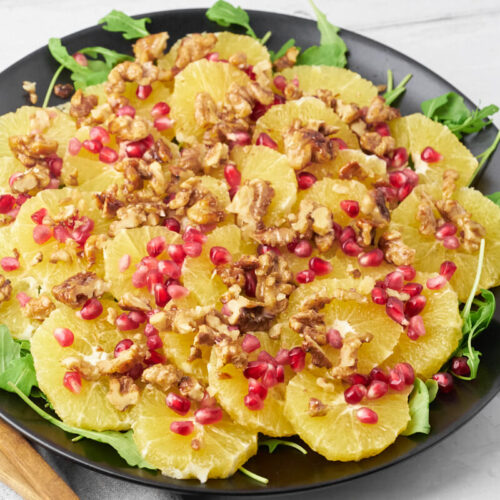 Orange salad with pomegranate and walnuts on a platter with salad utensils.