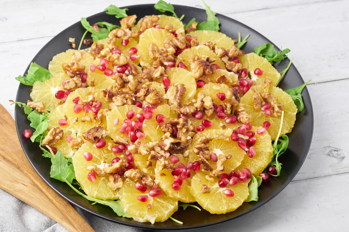 Orange salad with pomegranate and walnuts on a platter with salad utensils.